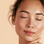 Beautiful asian girl model touching fresh glowing hydrated facial skin on beige background closeup