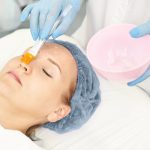 Woman undergoing a chemical peel treatment as a professional applies gel with a brush, highlighting skin rejuvenation, exfoliation, and wrinkle reduction.