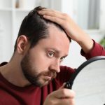 Man with hair loss problem looking at himself in the mirror indoors, considering treatment options like PRP therapy versus Minoxidil.